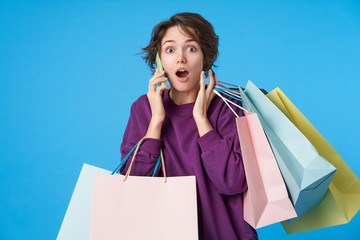 Wall Mural - Open-eyed young pretty dark haired curly lady raising amazedly her eyebrows while hearing surprising news on phone, standing over blue background with paper bags