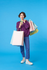 Wall Mural - Puzzled young pretty short haired curly female keeping mobile phone in raised hand and looking pensively upwards, posing over blue background with shopping bags