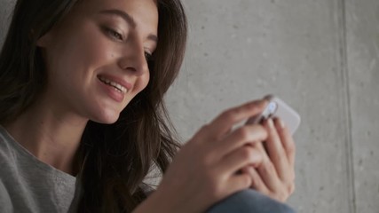 Wall Mural - Close up view of Smiling pretty brunette woman using smartphone and looking away after that while sitting near the wall at home