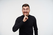 © timtimphoto - Portrait of excited young brown haired bearded man keeping raised forefinger on his mouth and raising surprisedly eyebrows while standing over white background