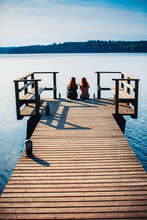 Two Girls Sitting  On The Edge Of The Wooden Pier Looking At The Calm Lake And Colorful Forest On The Other Side