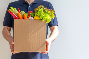 Food delivery service - man with a cardboard box of fresh vegetables on a gray background. Copy space