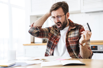 Wall Mural - Portrait of young shocked man holding credit card while working at home