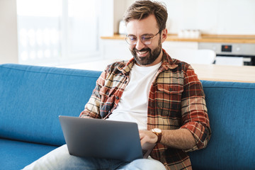 Canvas Print - Portrait of young happy man smiling and using laptop on sofa at home