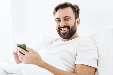 Canvas Print - Image of young unshaven caucasian man smiling and using cellphone while lying in bed at home