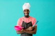 © Carlos David - Portrait of a smiling young male chef with arms crossed holding spatula and bowl, against blue studio background