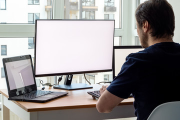 A man in a blue t-shirt is sitting at a workplace with two laptops and a monitor near the window. Remote work from home in quarantine