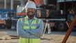 Portrait of Contractor / Investor / Architectural Engineer Wearing Hard Hat and Safety Vest Standing on a Commercial Building Construction Site, Crosses Arms Confidently. In Background Crane Machinery