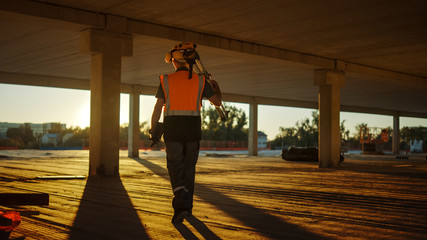 Wall Mural - Inside of the Commercial / Industrial Building Construction Site: Engineer Surveyor Finishes Day's Job Carries Tripod Theodolite and Walks into the Sunset. In Background Skyscraper Formwork Frames