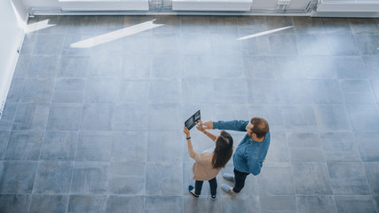 Wall Mural - Young Hipster Man and Female Stand in an Empty White Office and Map it with an Augmented Reality Software on a Tablet. Sunlight Shines Through Big Windows.