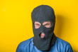 man close up thief in a mask and a blue shirt on a yellow background looks slyly to the camera. Mimicry. Gesture. photo Shoot/ evil criminal wearing balaclava