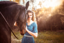 
Beautiful Girl With Her Wavy Hair In A Blue Dress Hugs A Horse And Smiles. A Bay Horse Stands In A Green Meadow In The Sunset. Magical, Fabulous Fantasy Photography.