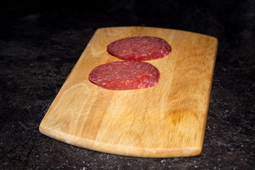 Wooden cutting board with two slices of cervelat on a dark table surface