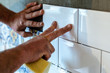 © Arseniy - Hands of a tiler man gluing a white rectangular ceramic tile on a gray wall. Repair work indoors.