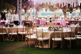 Fototapeta  - International Wedding outdoor celebration EVENING party under palm trees. Served tables on green area in hotel. Landyard. Beige and pink colors. Close-up and wide angle.