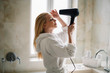 © JustLife - Young happy smiling woman wearing bathrobe using hair dryer in bathroom.