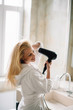 © JustLife - Young happy smiling woman wearing bathrobe using hair dryer in bathroom.