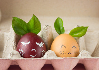 two chicken colored eggs with a cute painted face on them, lying in an egg box on a pink background, a festive Easter concept