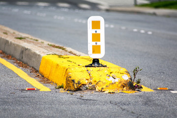 Dividing median curb on road with white and yellow warning reflecting sign on pole. Yellow traffic marking paint an overgrown vegetation