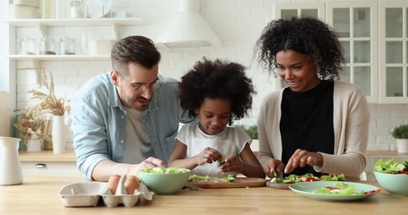 Wall Mural - Happy mixed race family of three young parents and afro american kid daughter enjoy cooking together. Multi ethnic mum and dad teaching cute child learning making healthy vegetable salad in kitchen.