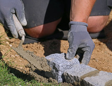 Close Up Photo Of Worker With Trowel And Gloves, Laying A Lawn Edge Massive Granite Blocks As A Part Of Garden Limitation. Detail Of Bricklayer's Working Hands Using Mortar