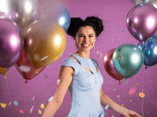 Portrait Of Young Woman With Black Curly Hair In Blue Party Outfit And Ballons Laughing And Celebrating Confetti Isolated On Pink Background