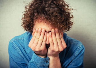 Curly young man crying, sad, covered his face with his hands. On a gray background