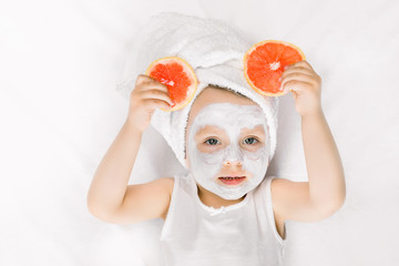 Wall Mural - Beauty, spa, hygiene concept for children. Cute little girl with towel on the head, white clay mask on face, posing to the camera and holding slices of grapefruit in her hands