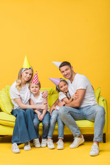 Wall Mural - cheerful family in birthday party caps hugging on sofa on yellow