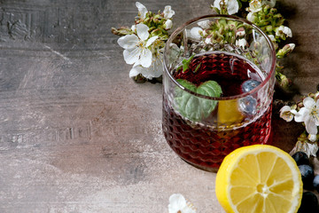 Wall Mural - Cocktail glass with blueberry sparkling cider or lemonade with sliced lemon, blueberries and mint. Blossom branches above. Gray texture background.