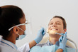 © LIGHTFIELD STUDIOS - selective focus of african american dentist in medical mask and face shield holding dental instruments near cheerful patient