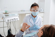© LIGHTFIELD STUDIOS - selective focus of african american dentist in medical mask and face shield near patient in clinic
