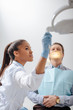 © LIGHTFIELD STUDIOS - selective focus of beautiful african american dentist in latex glove touching medical lamp near patient