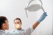 © LIGHTFIELD STUDIOS - selective focus of attractive african american dentist in latex glove touching medical lamp near patient