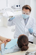 © LIGHTFIELD STUDIOS - selective focus of dentist in face shield, medical mask and latex gloves touching medical lamp near african american woman