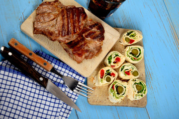 two pieces of grilled meat on a cooking plate with pieces of tortilla next to a cold drink on an old blue table