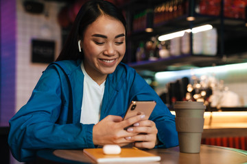 Photo of asian woman using mobile phone and wireless earphones in cafe