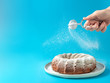 canvas print picture - Woman's hand sprinkling icing sugar over fresh home made bundt cake. Powder sugar falls on fresh perfect bunt cake over blue background. Copy space for text. Ideas and recipes for breakfast or dessert