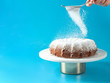 © fascinadora - Woman's hand sprinkling icing sugar over fresh home made bundt cake. Powder sugar falls on fresh perfect bunt cake over blue background. Copy space for text. Ideas and recipes for breakfast or dessert