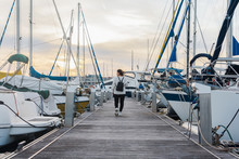 Back View Of Faceless Young Lady In Casual Wear With Black Backpack Walking On Wooden Pier Between Modern Yachts On Sunset
