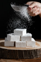 Crop Person With Sieve Sprinkling Sugar Powder Over Pieces Of Marshmallow Placed On Wooden Board Against Black Background