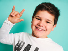 Joyful Confident Chubby Plus Size Preteen Boy In Casual Clothes Looking At Camera And Laughing While Making Rock And Roll Sign Against Turquoise Background