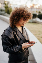 Young Businesswoman In Retro Black Leather Jacket And Skirt Browsing Smartphone While Standing In Arched Passage In Park Before Work