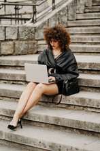 Clever Curly Businesswoman Wearing Black Leather Suit And Jacket Typing On Laptop Keyboard While Sitting On Stairs And Working On Remote Project On City Street