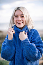 Joyful Cheerful Young Blonde Female In Blue Knitted Sweater Looking At Camera And Laughing While Standing Against Blurred Background Of Sea Coast