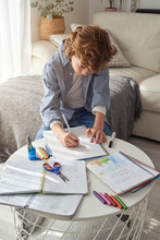 High Angle Of Boy With Curly Hair Sitting On Sofa Near Round Table And Writing In Notebook While Doing Homework In Cozy Living Room At Home