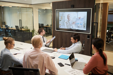 Mature businesswoman o the screen holding the online conference for business people who sitting at the table and listening to her at office