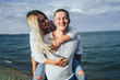 © Daria - Attractive blonde girl jumped onto her boyfriend's back. He carries her, she laughs and kisses him. They run along the sand on the seashore on a summer evening, lit by the sun. Romantic Date Concept