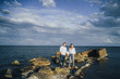 © Daria - Long shot. Attractive couple in bright clothes standing on stones in the sea against a blue cloudy sky. Lit by the soft evening sun. Looking forward, hug, kissing and enjoying beautiful evening