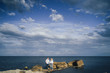 © Daria - Long shot. Attractive couple in bright clothes standing on stones in the sea against a blue cloudy sky. Lit by the soft evening sun. Looking forward, hug, kissing and enjoying beautiful evening